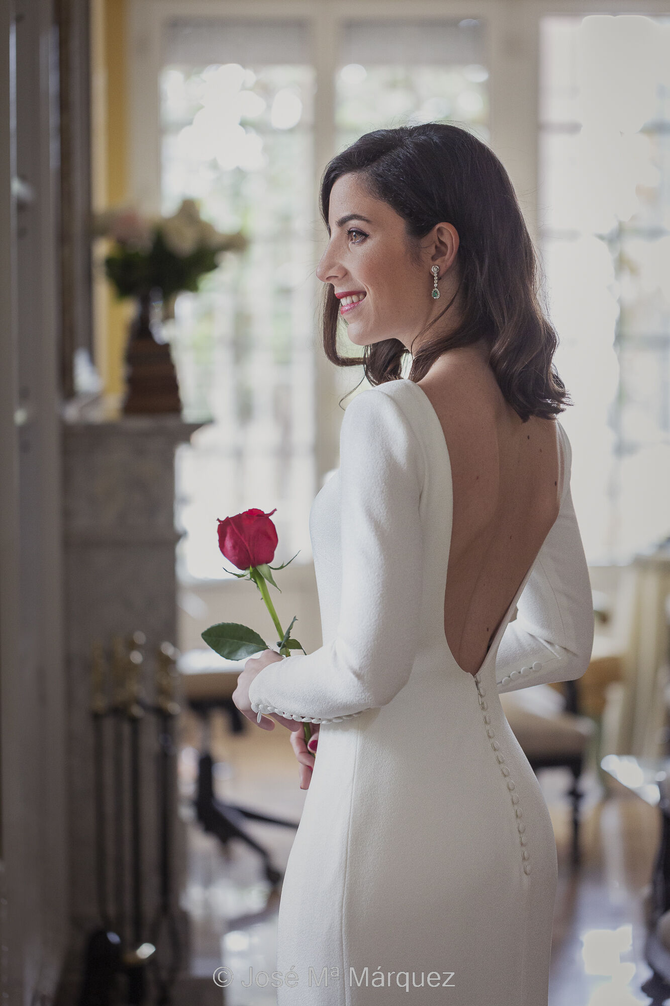 Retrato de la novia ya vestida y preparada en el salón de su casa, justo antes de salir hacia la ceremonia. Es un retrato de perfil, de medio cuerpo, iluminado por la luz de una gran ventana y con otra gran ventana como fondo. Fotografía de boda en Granada. Fotógrafo José María Márquez. Fotografía de boda. Retrato de una novia. Foto vertical y en color. La novia aparece en el salón de su casa ya vestida y preparada justo antes de salir hacia la ceremonia. Encuadre en plano medio. Está de pie, de perfil mirando hacia la izquierda. Se intuye que mira fuera a través de una gran ventana, que es la que le ilumina suavemente, casi de forma envolvente, sin sombras pronunciadas. La cara de la novia mira hacia la ventana pero su cuerpo está ligeramente girado hacia su derecha, de forma que deja ver el vestido por atrás, luciendo la espalda al aire y los botones del vestido hacia abajo, dibujando una esbelta y atractiva figura femenina. Sostiene delicadamente una rosa roja entre sus manos y sonríe levemente. Al fondo, muy desenfocados, se ven una chimenea de mármol y un gran ventanal. Es una foto tomada el día de la boda por José Mª Márquez, fotógrafo de Granada.