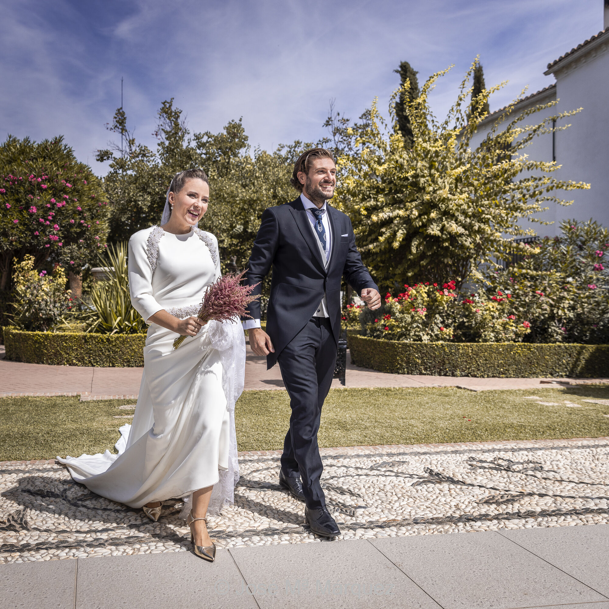 Fotografía instantánea de los novios entrando al cóctel. La pareja camina hacia los invitados sonriendo. Fotoperiodismo de boda. Documentalismo estético. Fotógrafo Márquez de Granada. Una pareja camina ufana y feliz en dirección a la derecha del fotógrafo, a donde se intuye que están los invitados de la boda. Los novios llegan en ese momento al cóctel al aire libre del patio ajardinado del restaurante, con sol de medio día. Es una instantánea cuadrada en color que documenta la alegría de los novios al encontrarse con sus amigos e invitados. La expresión corporal de ambos en el movimiento detenido es estética, con mucha plasticidad y real, sin pose, coherente con la felicidad del momento y con el estilo fotoperiodístico de boda del fotógrafo de Granada José Mª Márquez.