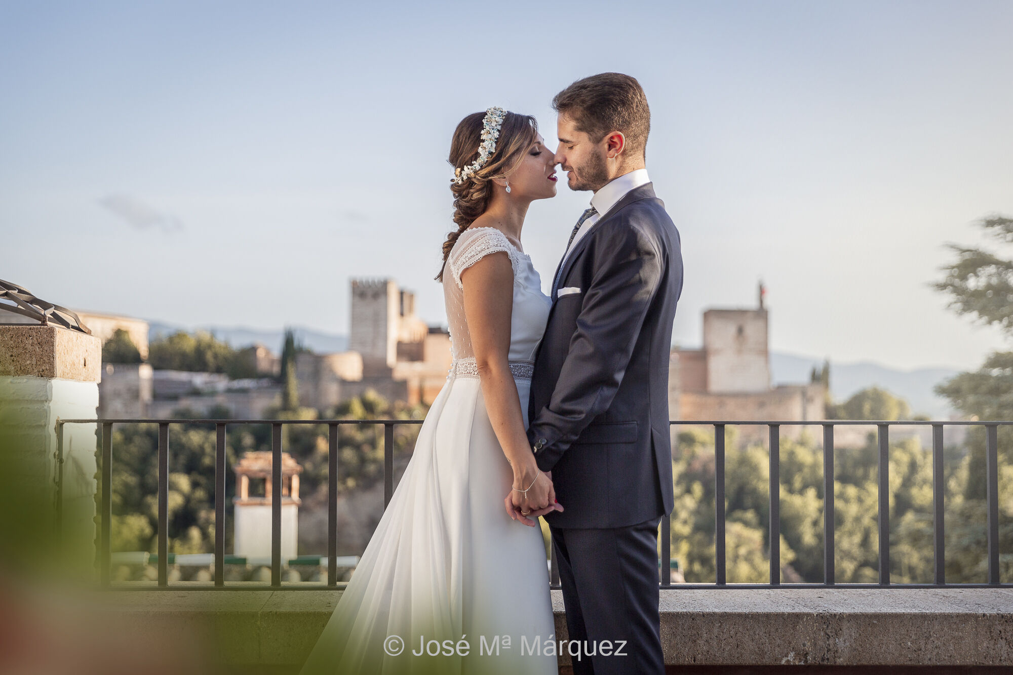 Una pareja de novios en su reportaje postboda en la mezquita del barrio del Albaicín (Granada) Están cogidos de la mano, frente a frente con las caras muy juntas, casi besándose, mirándose a los ojos. La foto está tomada por el fotógrafo de bodas de Granada José Mª Márquez. Justo detrás de ellos está la baranda del mirador y al fondo la Alhambra, iluminada lateralmente por la luz del atardecer. Fotógrafos Granada.