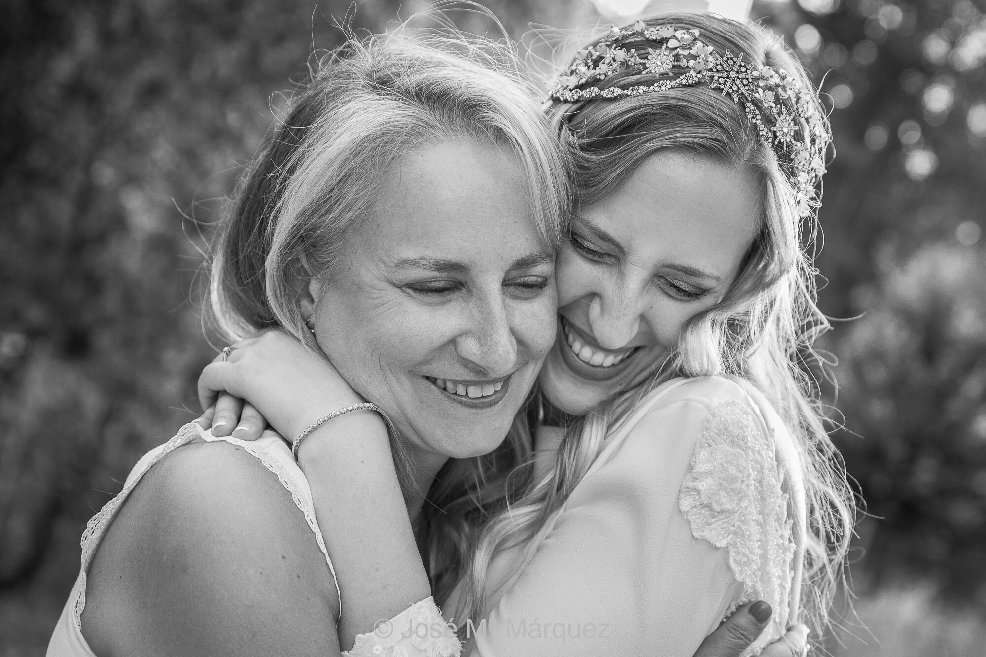 La novia y su madre bromean y se abrazan en un momento del reportaje de postboda. Primer plano de una escena tierna y llena de amor. La complicidad y la cercanía en el trato hace posible que el fotógrafo capte estas instantáneas con expresiones tan auténticas. José Mª Márquez, fotógrafo de Granada. Fotografía de Boda. Horizontal. Blanco y negro. Plano corto de una novia con su madre encuadradas desde los hombros. Madre e hija se abrazan y sonríen tiernamente con las caras juntas en un esto cariñoso y tierno. La cercanía en el trato del fotógrafo durante el reportaje hace posible captar instantáneas donde la cercanía emocional de los protagonistas se evidencia de forma natural. Es un ejemplo de fotografía documental estética del fotógrafo de boda de Granada José María Márquez.