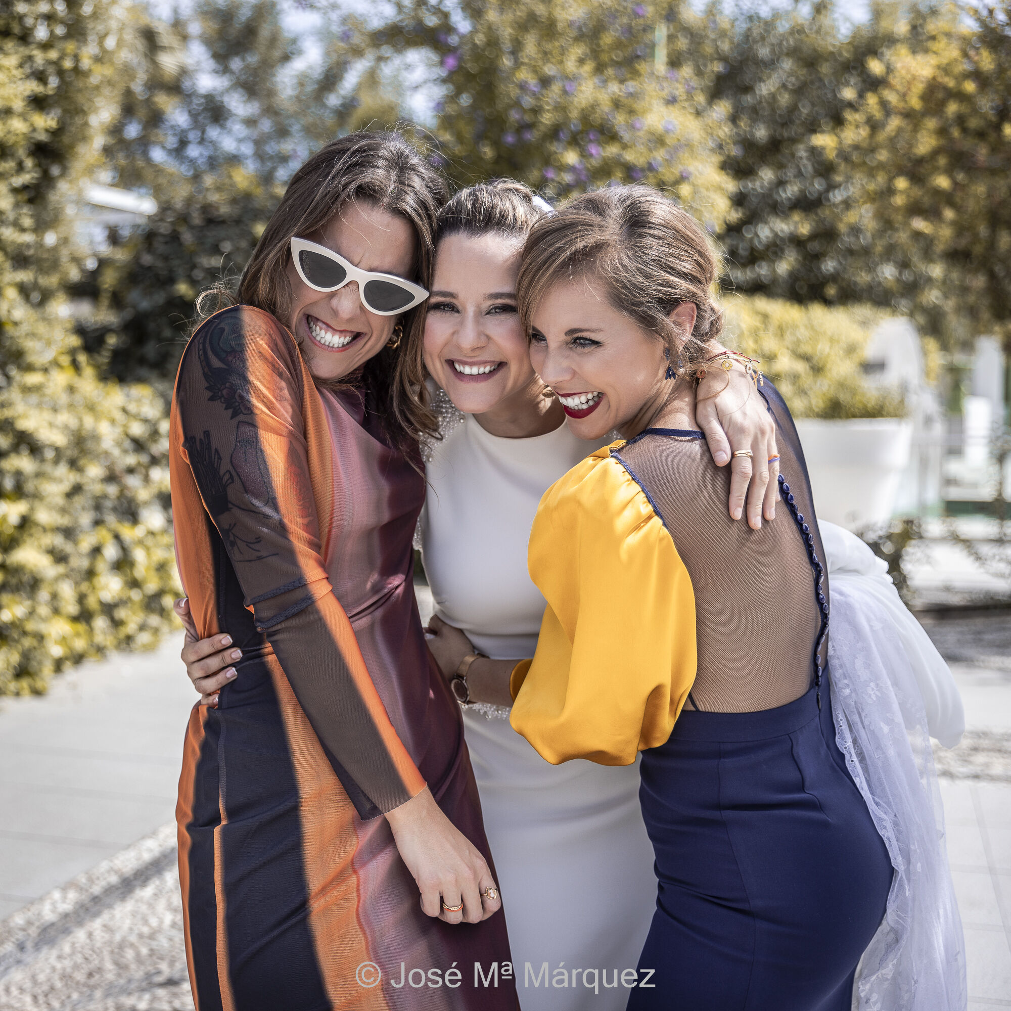 Foto periodismo gráfico de boda. Instantánea que documenta un momento divertido en el que dos amigas abrazan a la novia y ríen espontáneamente durante el cóctel. Fotógrafo de bodas JM Márquez, Granada. Instantánea de boda cuadrada y a color. Cóctel de boda al aire libre, con luz de mediodía, en el patio ajardinado de un salón de bodas cerca de Granada. La foto documenta un momento divertido en el que dos amigas, una a cada lado de la novia, la abrazan. Las tres ríen espontáneamente giradas levemente hacia el fotógrafo. Documentalismo estético de boda. Fotógrafo José María Márquez.