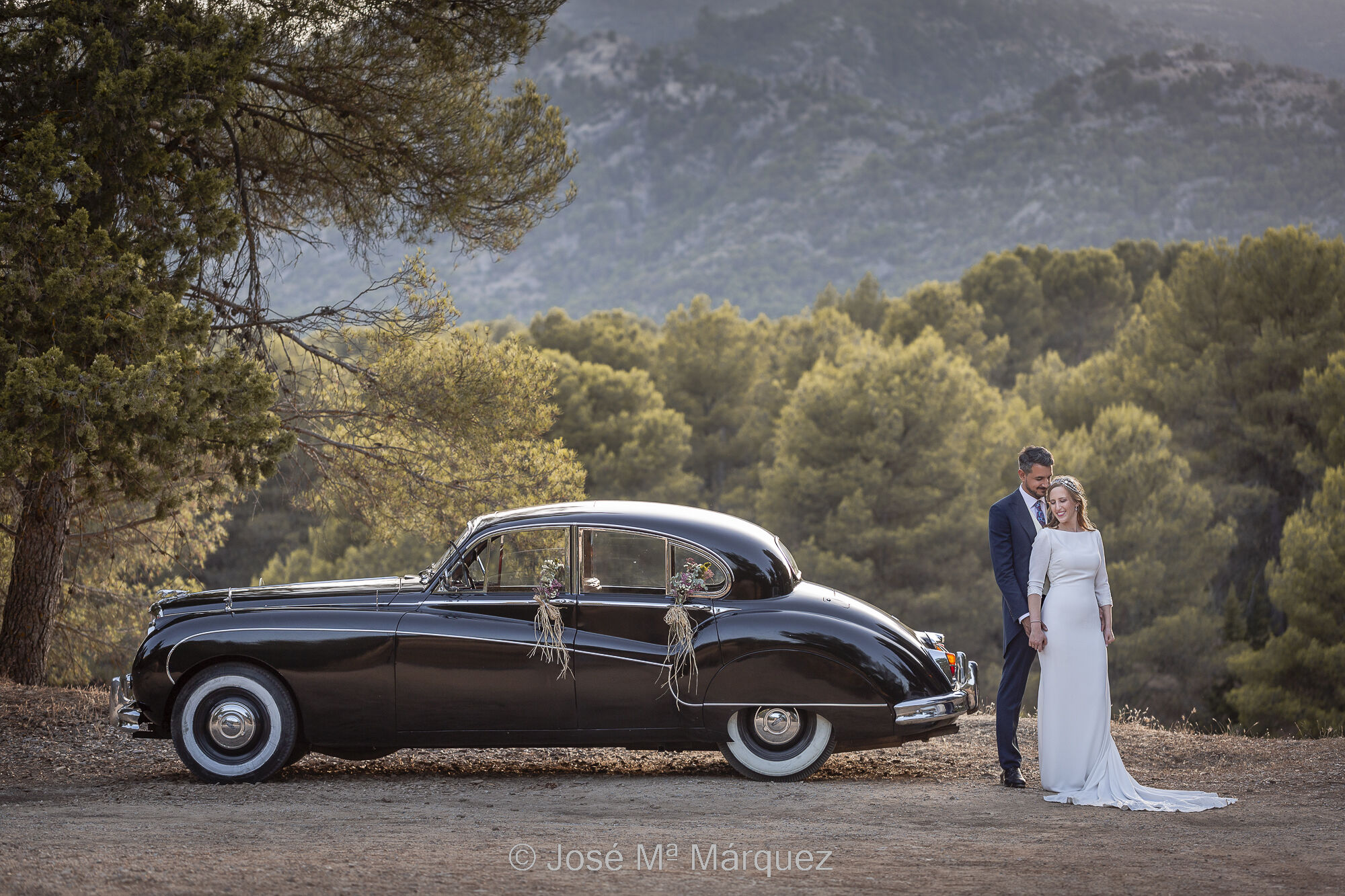 Foto del reportaje de boda de unos novios por la sierra de Cazorla durante la sesión de exteriores, entre la ceremonia y el cóctel. Los novios están al filo de un cortado de pié junto a un coche de época negro adornado con flores. La luz de contra de la tarde siluetea a la pareja y al coche. Al fondo se ve un paisaje arbolado muy espeso iluminado también con esa luz de contra que da un bonito volumen a las copas de los árboles. El novio, más alto, está detrás de la novia y le coge las menos, mientras ella gira su cabeza hacia el pecho de él con un gesto relajado. Foto de José Mª Márquez. Fotógrafos de Granada.