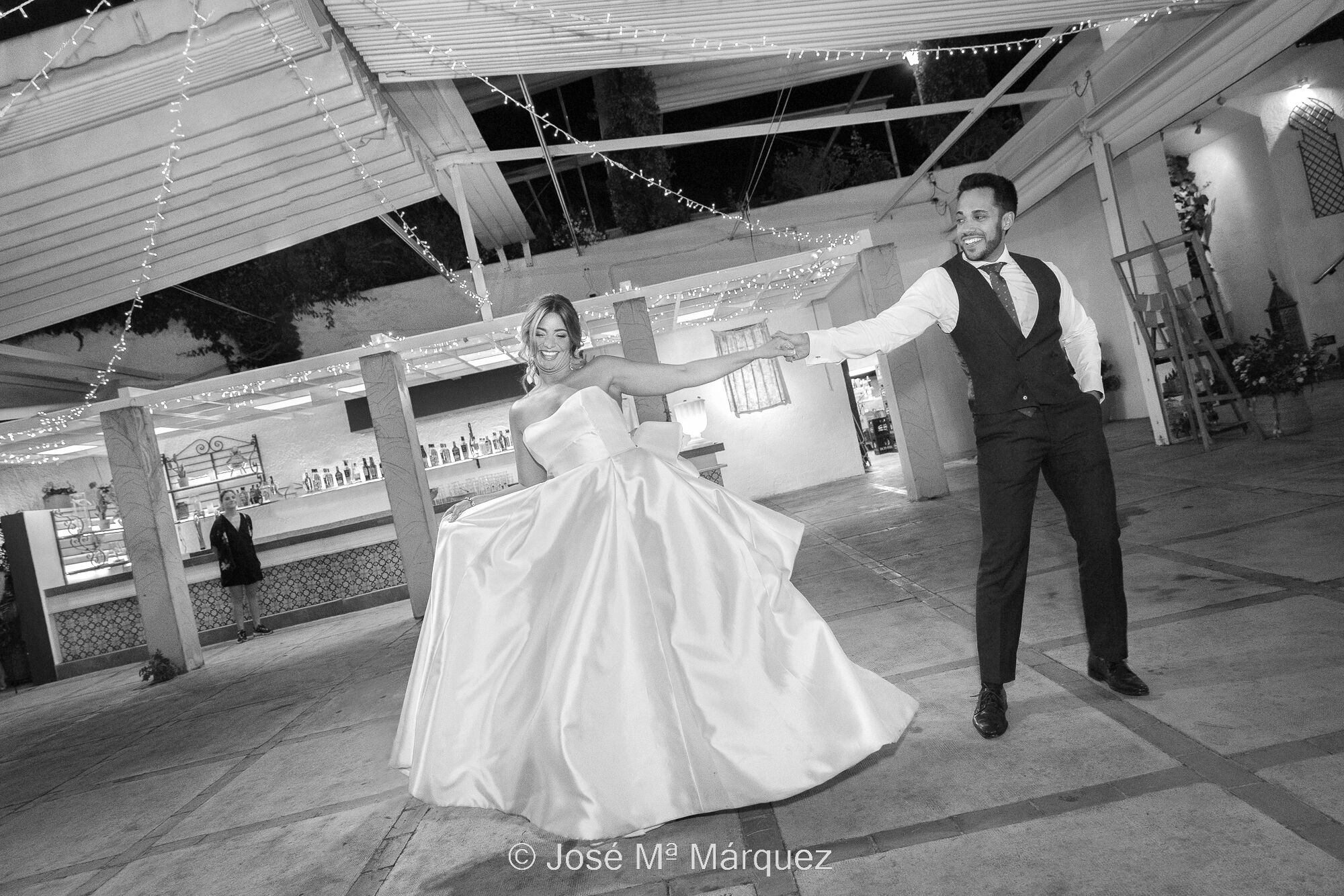/content/img/gal/3052/pareja-recien-casada-inaugura-el-baile-en-la-celebracion-boda-en-la-chumbera-sacromonte-baile-de-los-novios-fotografos-boda-granada.jpg Foto horizontal y blanco y negro. Instantánea de boda. Una pareja inaugura el baile en la recepción celebrada en La Chumbera (barrio del Sacromonte), Granada. Los novios están cogidos de la mano frente a la cámara en un gesto detenido de la coreografía con expresión simpática y elegante. Cada uno tira hacia su lado, manteniendo estirados sus brazos. El con su mano izquierda en el bolsillo y ella sosteniendo con su derecha el vuelo del vestido con gracia. El fondo se ve la barra del bar y la encargada del restaurante. Estilo de trabajo: Fotoperiodismo (documentalismo) estético de boda. Autor: El Fotógrafo de Bodas de Granada JM Márquez.