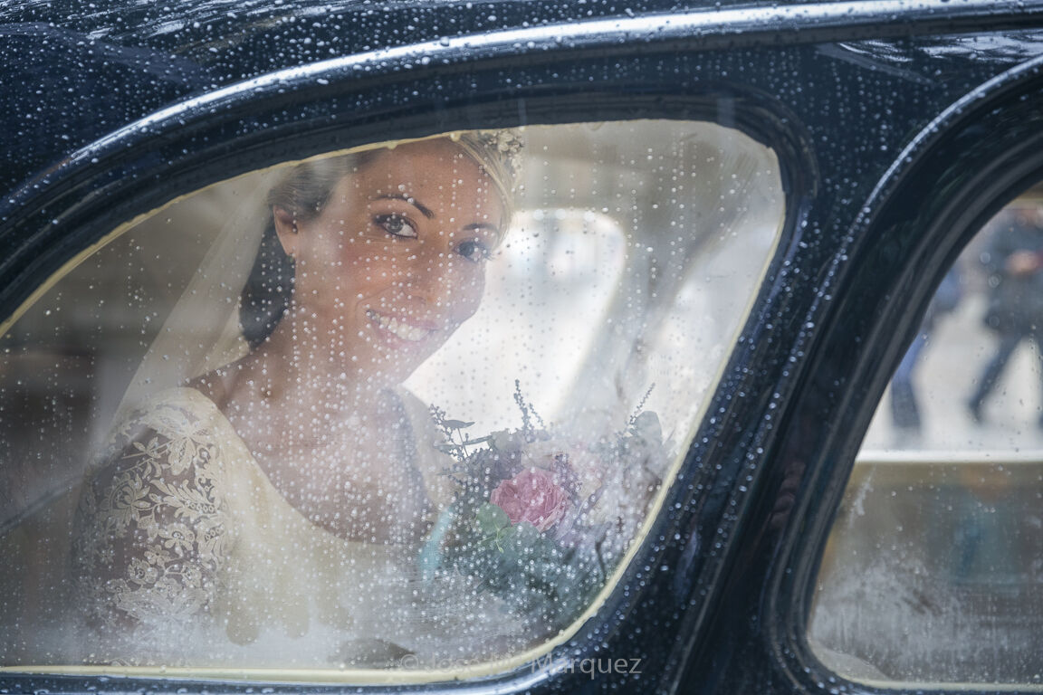 Día de boda lluvioso. Retrato de la novia a través de la ventanilla trasera del coche nupcial en la llegada a la Iglesia. El cristal lleno de gotas aporta belleza y narrativa a la imagen. Fotografía de boda del fotógrafo granadino José María Márquez. Fotografía de boda horizontal y en color. Día de boda lluvioso. El encuadre coincide con la ventanilla trasera del coche nupcial a la llegada a la iglesia. Es un bello retrato de la novia a través del cristal lleno de gotas de lluvia. La escena tiene un aire melancólico. La novia sonríe mirando a cámara sosteniendo su ramo. Es un ejemplo perfecto de narrativa en fotografía de boda, donde unos pocos elementos gráficos aportan información fundamental para contextualizar el momento. Es una foto de José Mª Márquez, fotógrafo de bodas en Granada.