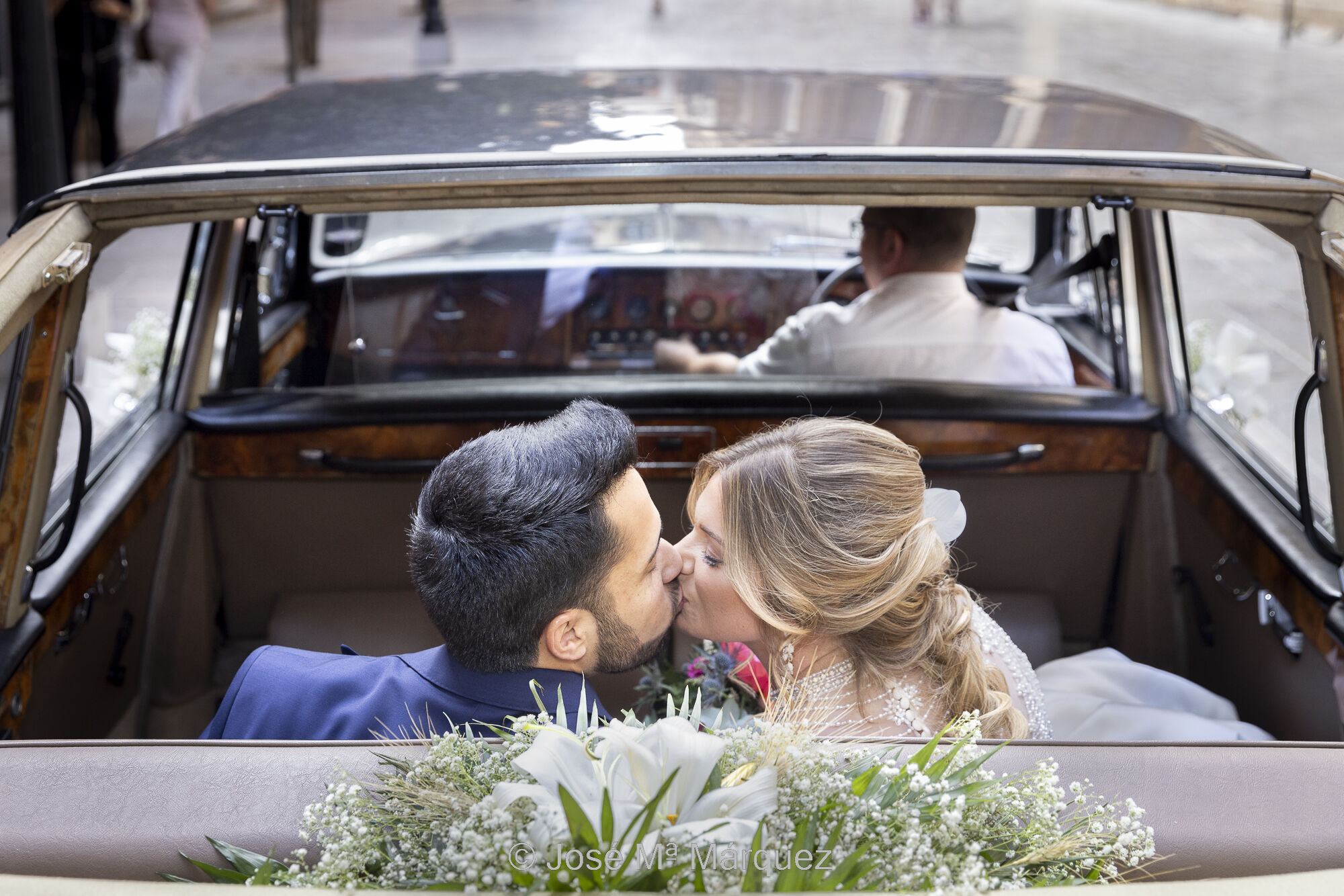 Foto del reportaje de bodas de una pareja en un coche de época descapotable. La foto está tomada desde la parte trasera y un poco elevada. Los novios están de espaldas a la cámara y se giran el uno al otro para besarse. Al fondo de ellos se ve el interior del vehículo, el chófer y gente desenfocada que va por la calle. El autor es José Mª Márquez, fotógrafo de bodas en Granada. 