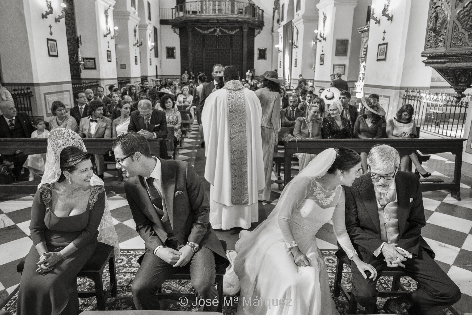 Momento de la ceremonia de boda en San Pedro en el que suceden tres historias simultáneamente. Mientras que el sacerdote da la comunión a los invitados, la novia departe con el padrino y el novio con la madrina. Son tres fotos dentro de una misma imagen. Fotografía de reportaje de boda. Horizontal, blanco y negro. Durante la ceremonia religiosa en la Iglesia de Granada de San Pedro y San Pablo. Punto de vista de la toma desde el altar, hacia los novios, padrinos y todos los asistentes de la boda, composición simétrica. Aparecen los novios y los padrinos en primer término sentados. El sacerdote está detrás de ellos, de espaldas a cámara, (en la imagen entre el novio y la novia), dando la comunión a los invitados, en el centro de la imagen. A un lado y a otro, simétricamente y cada uno por su lado, la novia, a la izquierda de la imagen, habla con su madre y el novio, a la derecha, departe tranquilamente con su padre. Es un ejemplo de fotografía de boda donde se dan varias pequeñas historia sincronizadas; una instantánea donde la narrativa y la composición son muy claras. Fotografía tomada por el fotoperiodista de boda granadino José Mª Márquez.