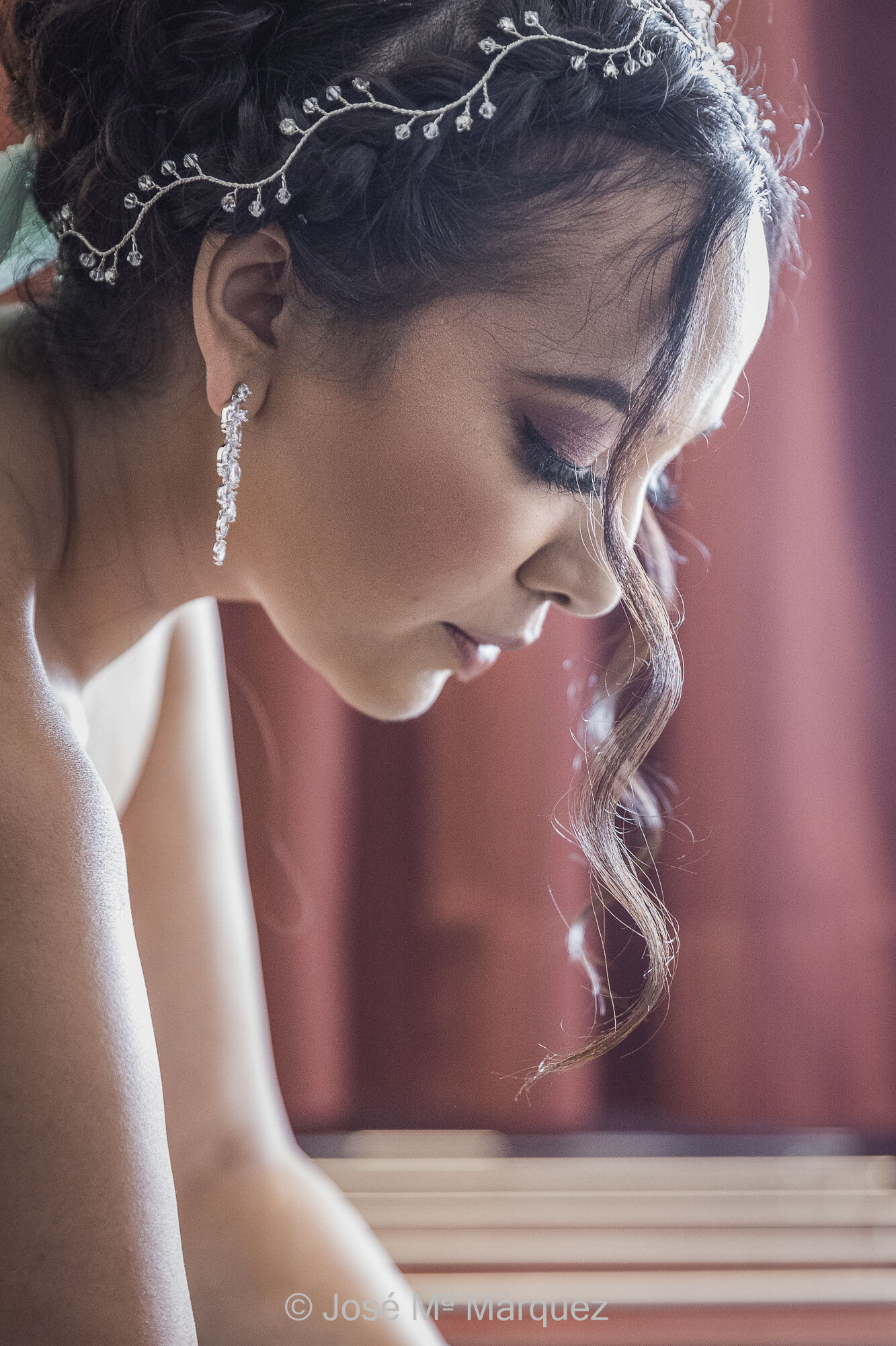 Retrato de Boda. Perfil. Primer plano de una bella novia hindú iluminada con luz natural lateral para acentuar los rasgos de la silueta. Fotógrafos boda Granada. Fotógrafo Márquez. Retrato de perfil de una bella novia durante sus preparativos. Foto vertical a color. Es un primer plano en el que la novia mira hacia abajo con la cabeza inclinada hacia la luz, que entra desde atrás y por la derecha, de modo que siluetea todo contorno (pestañas, ojos y boca). Dos mechones del peinado le cuelgan desde su frente por delante de la cara. Una diadema y unos largos pendientes plateados adornan su rostro. Se ha usado como fondo una cortina granate desenfocada. Se ha usado como fuente de iluminación la ventana de la habitación del hotel. La fotografía se tomó por el fotógrafo de bodas de Granada José María Márquez.