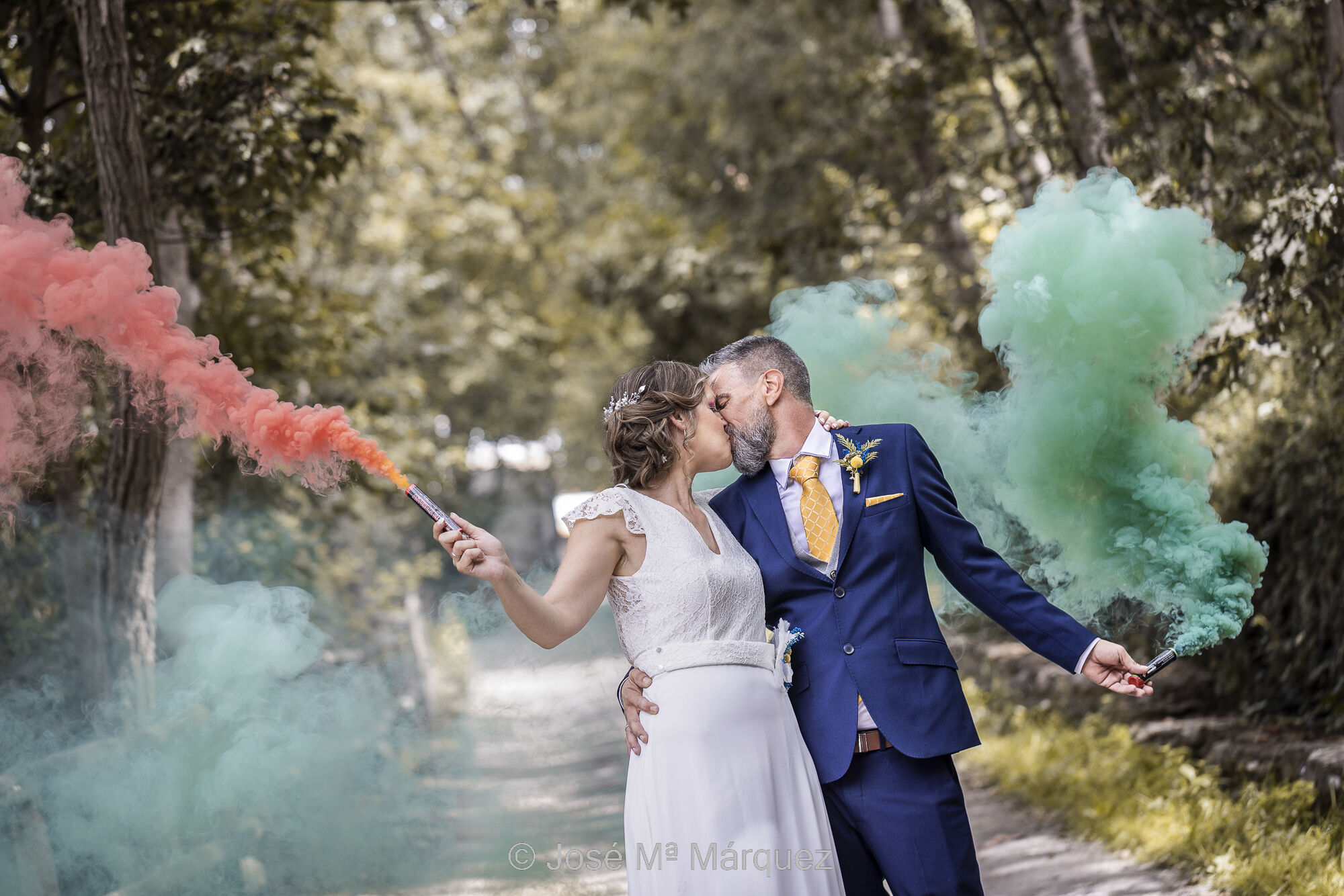 Una pareja pasea con bengalas de humo. Los novios se han detenido y se besan mientras el humo rojo y verde se extiende por el fondo. Fotografía de boda con plasticidad compositiva. Foto de boda del fotógrafo de Granada José María Márquez. Fotografía de boda. Horizontal. Foto en color. Plano medio de una pareja paseando por un camino arbolado en la sesión de exteriores. Detrás de ellos se ve desenfocado el camino en el bosque por el que vienen. Encuadre por debajo de la cintura. Cada uno lleva una bengala de humo de colores. Ella, a la izquierda de la foto, la lleva en su mano derecha y él en la izquierda. Con la otra mano se rodean por la cintura, de cara a la cámara, mientras se besan en los labios. Se ve un gesto natural y la instantánea luce con plasticidad compositiva por la expresión corporal de los novios. Fotografía de José María Márquez, fotógrafo de bodas de Granada.