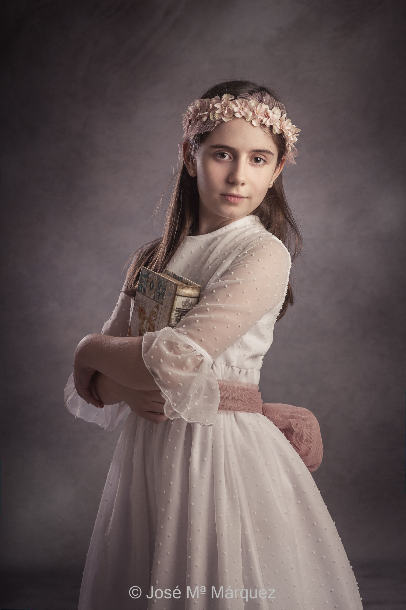 Foto de Estudio de una niña con su vestida de primera comunión con los brazos cruzados y un libro entre ellos. Autor José María Márquez, fotógrafo de primeras comuniones en Granada. Estudio Fotográfico