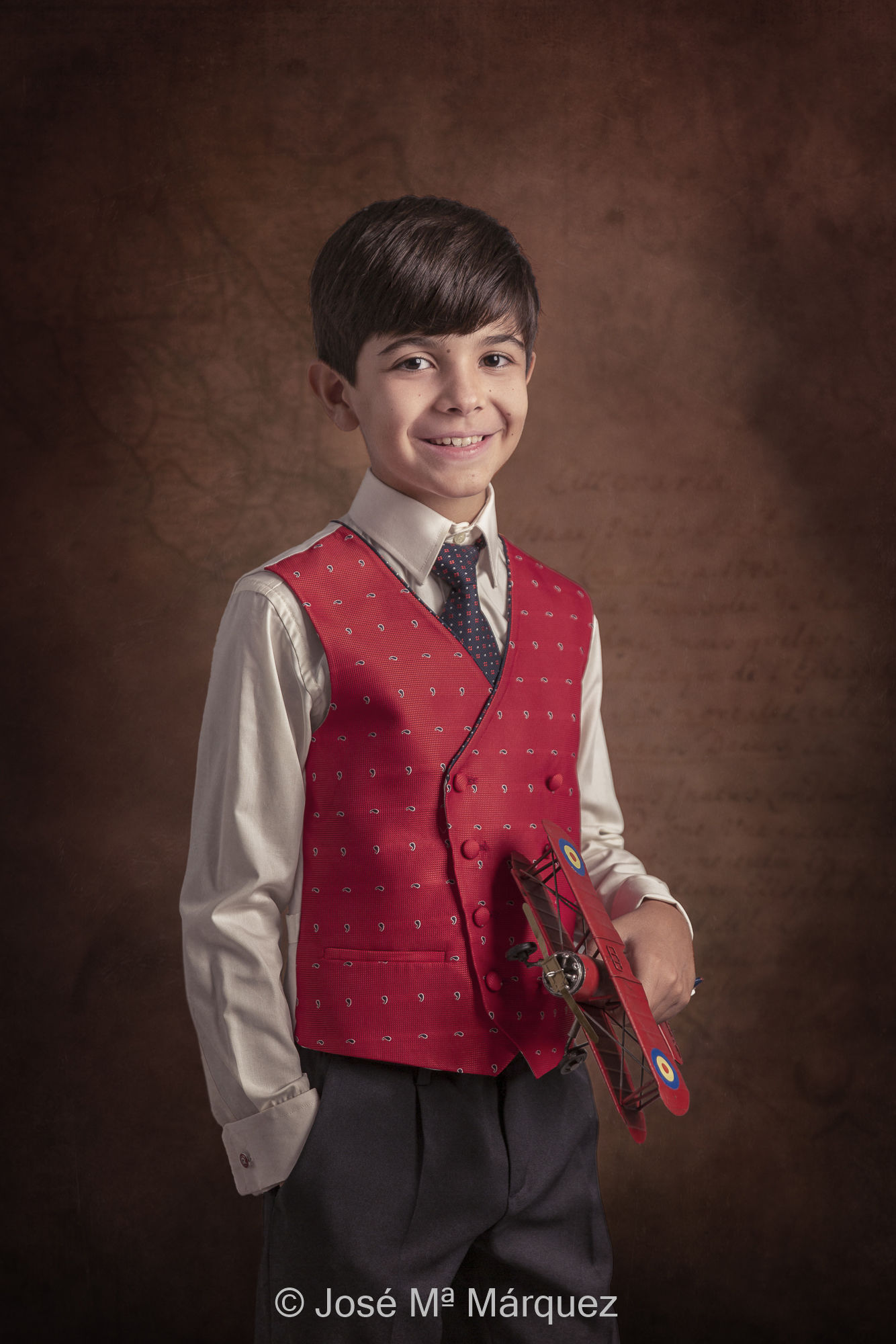 Fotografía de Estudio de un niño con un chaleco rojo de su traje de primera comunión y un avión antiguo en la mano. Fotógrafo de Granada José Mª Märquez