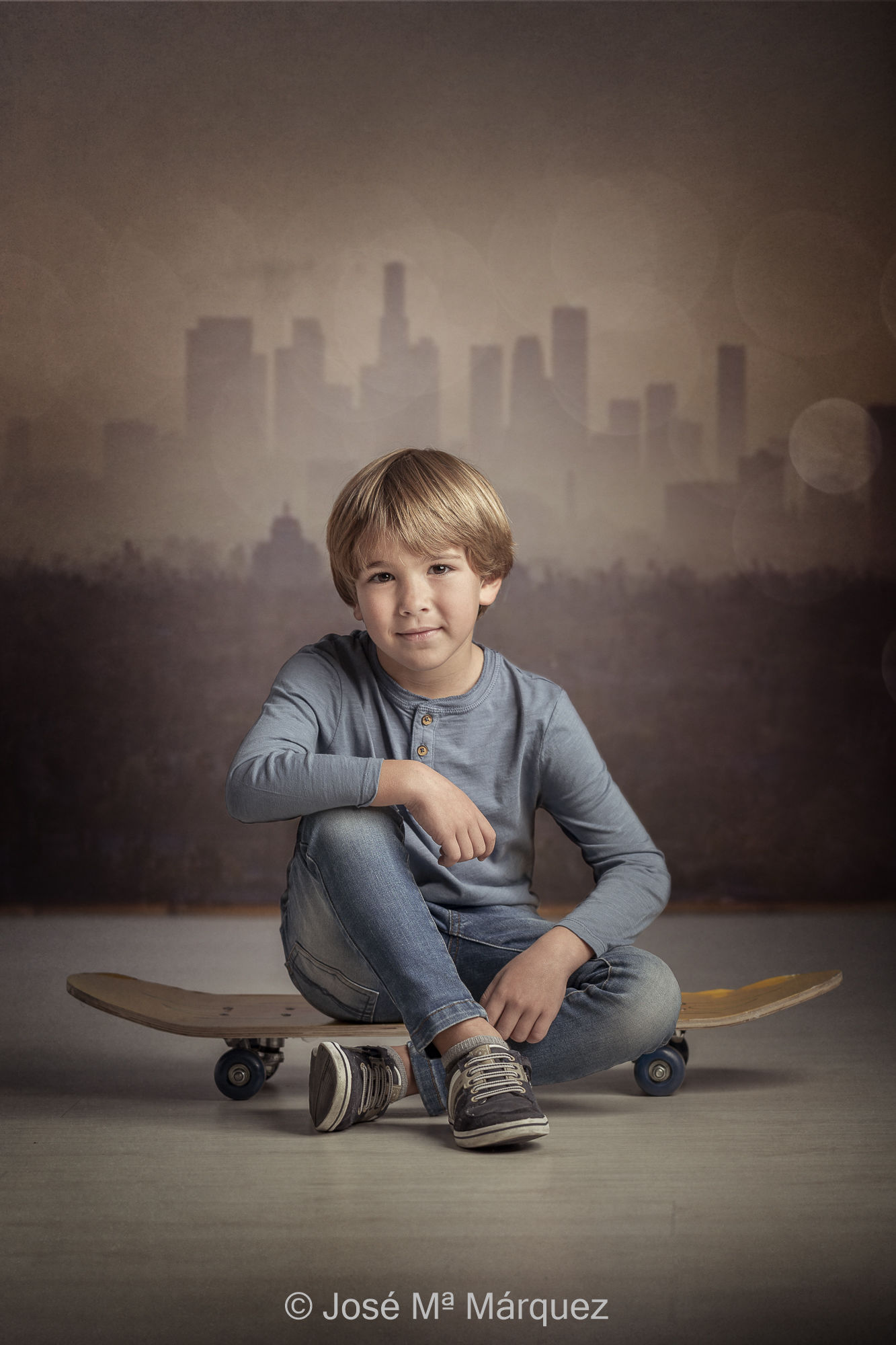 Fotografía infantil escenificada con temática urbana. Niño sentado en monopatín con skyline al fondo