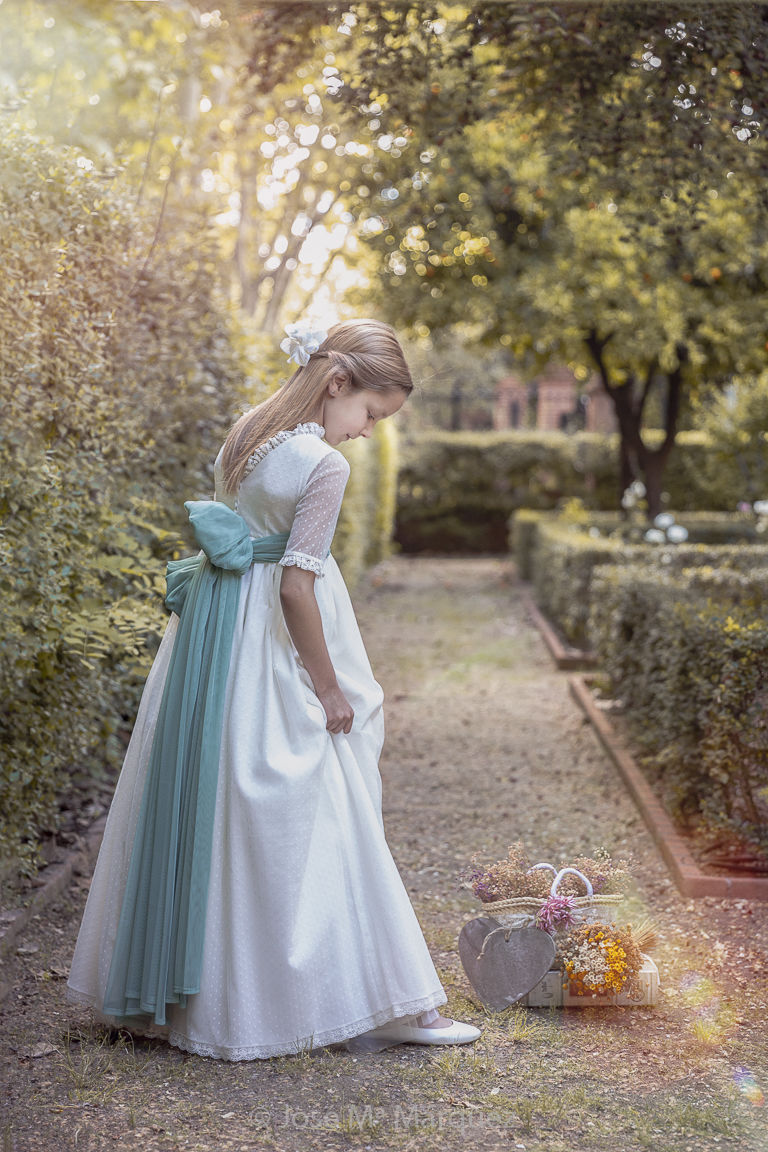 Foto en exteriores de una niña de cuerpo entero vestida de primera comunión. La luz natural de contra, el boqué, el balance cromático y la pose delicada crean una escena etérea y dulce muy romántica y apropiada para el género de Retrato de Comunión. Autor: José Mª Márquez, estudio fotográfico de Granada