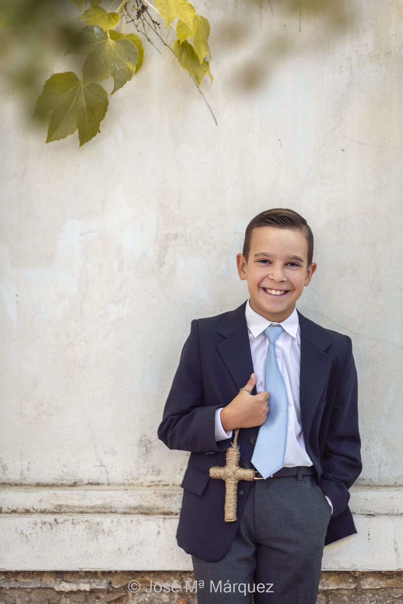 Sesión en exteriores de Primera Comunión. El niño con traje de chaqueta y corbata azul posa feliz  apoyado en la pared del jardín sujetando una cruz de cuerda y ríe. Fotógrafo José Mª Márquez. Estudio fotográfico de Granada. Fotografía de un reportaje de comunión.