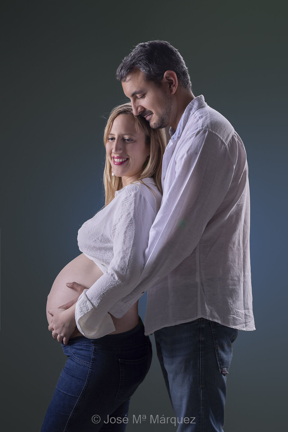 Sesión de embarazo en estudio. El chico abraza desde atrás a su mujer embarazada rodeando el vientre con sus brazos.