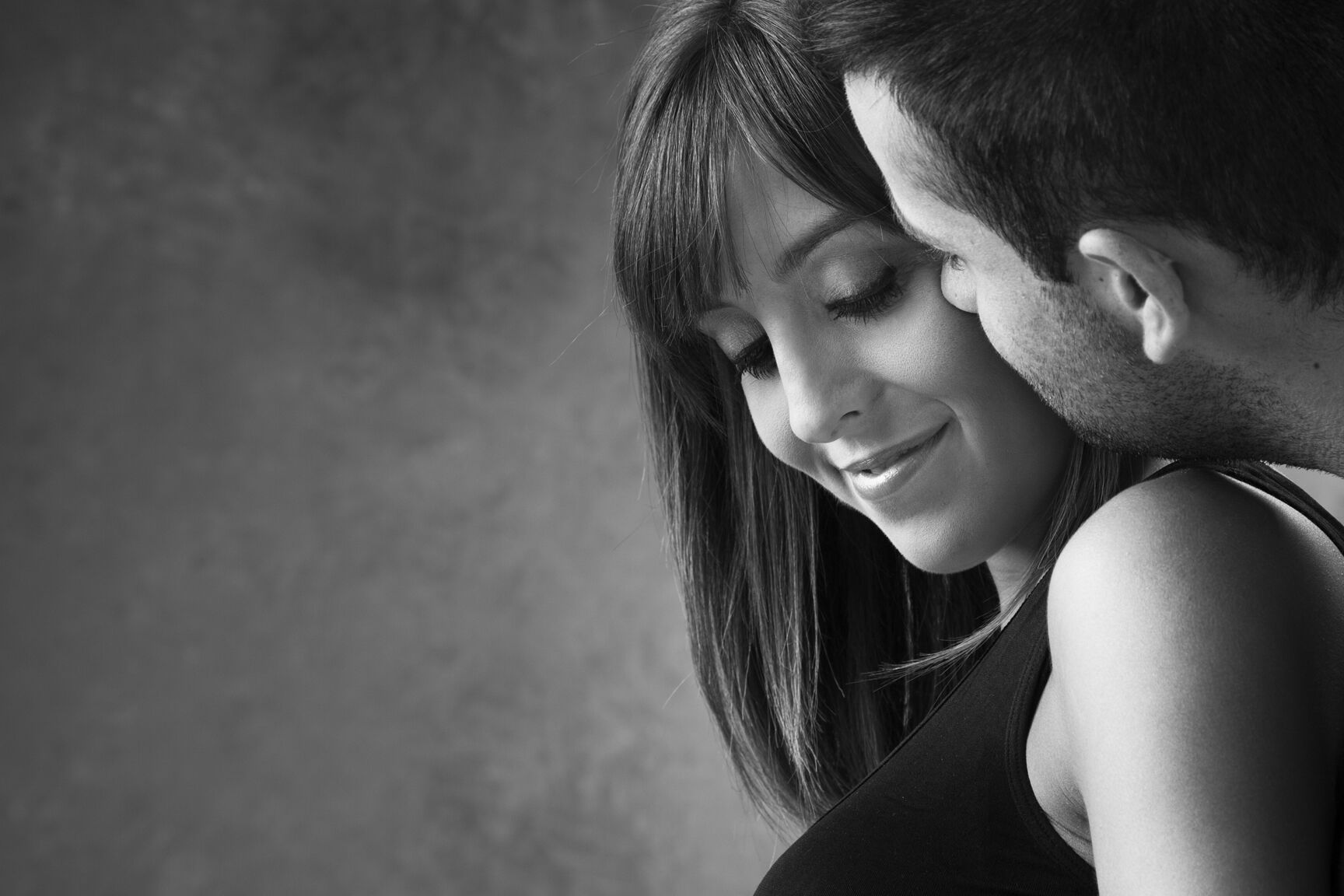 Retrato en blanco y negro de una sesión de pareja en el estudio. El futuro papá besa tiernamente a su esposa embarazada en la mejilla.