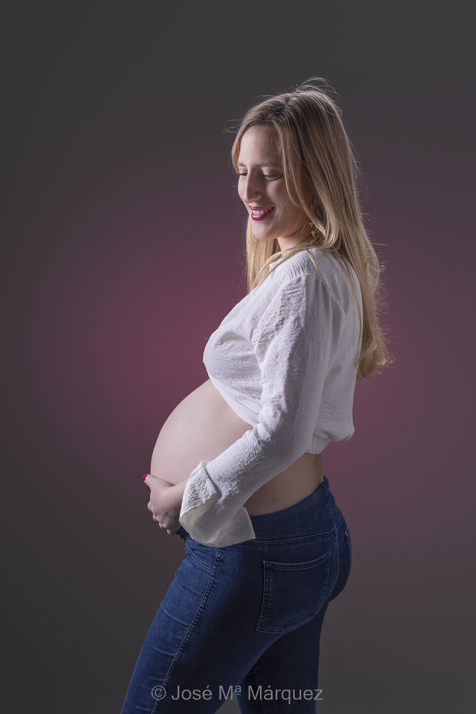 Foto de una chica embarazada en el estudio Plano medio de una embarazada con iluminación clásica en el estudio. La chica vestida con vaqueros y blusa blanca se acaricia el vientre.