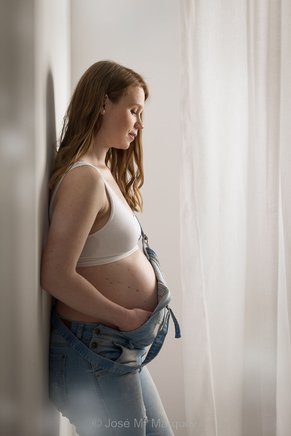 Foto premamá de una chica en el estudio La futura mamá viste con un peto vaquero y top blanco. Se acaricia el vientre por debajo del peto. Fotografía de embarazo en estudio.