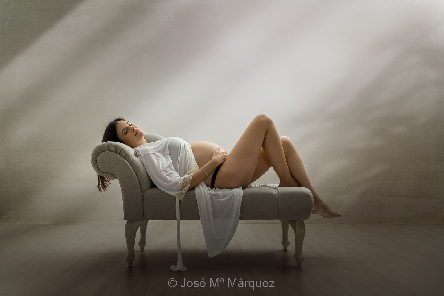 Retrato en estudio de una chica embarazada. La mujer está recostada bocarriba en un cheslón con los ojos cerrados, llevando una bata blanca que cae hasta el suelo. La amplia luz cenital crea un suave volumen y un ambiente dulce y plácido.