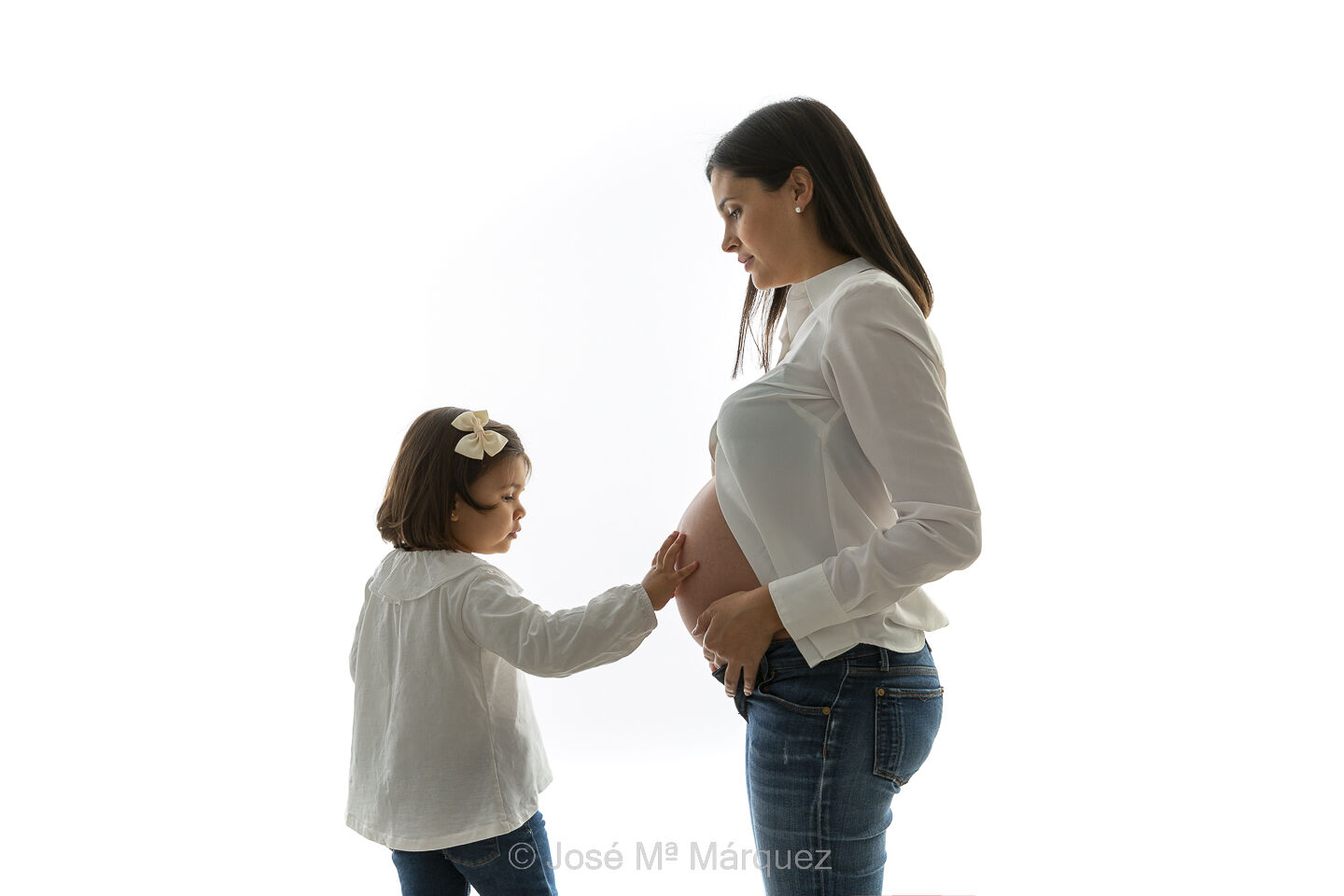 Fotografía de embarazo en estudio iluminada en clave alta con luz de rebotada de contra. La hija toca suavemente el vientre de mamá.