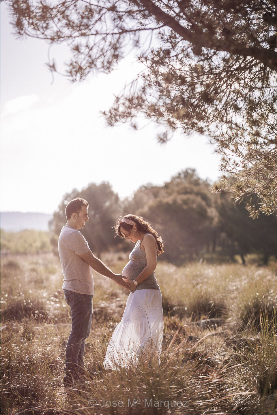 Pareja embarazada en el campo. Plano general. Foto de una sesión de embarazo de exteriores en el campo de una pareja. Plano general vertical de perfil con luz de contra. El chico está frente a su mujer alargando la mano hasta el vientre.