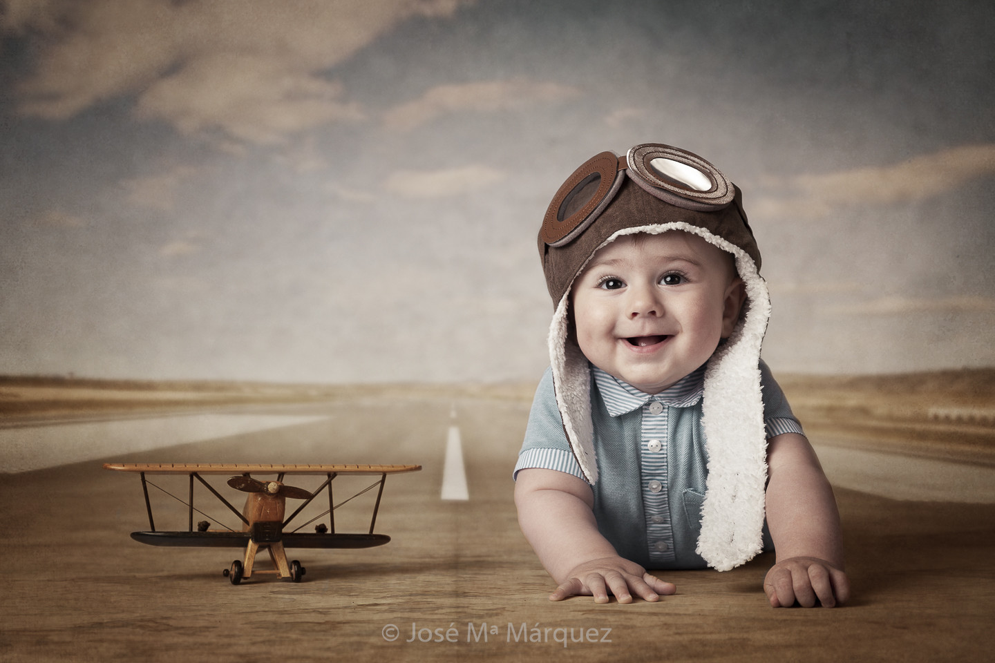 Bebé con gorrito de aviador y avión clásico en la pista de aterrizaje. Sesión fotográfica en estudio de José María Márquez. Fotografía Infantil